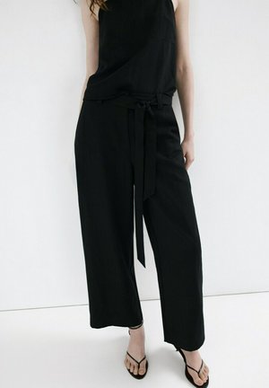 Mujer con top negro sin mangas y pantalones de pierna ancha con cinturón de tela, combinados con sandalias negras de tacón abierto, de pie frente a una pared blanca.