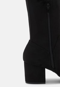 Černá semišová kotníčková bota s blokovým podpatkem a viditelným bočním zipem na bílém pozadí.