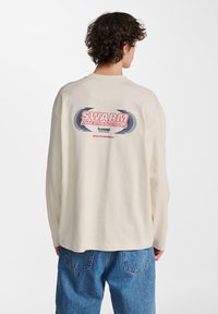 Cremfarbendes Langarmshirt mit einem großen "SWARM INTERNATIONALS"-Grafik in Rot und Blau auf dem Rücken, aus weichem Baumwollstoff gefertigt.