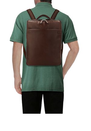 Homme portant un polo vert et un pantalon noir avec un sac à dos rectangulaire en cuir marron foncé sur le dos, vu de dos.