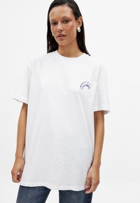 T-shirt en coton blanc avec une petite broderie de croissant bleu sur la poitrine gauche. Coupe ample avec des manches courtes et un col rond.