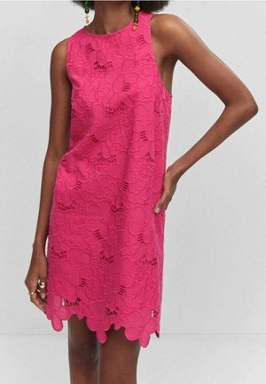 Robe sans manches en dentelle rose vif avec de grands motifs floraux et un ourlet festonné portée par une personne avec une main sur la hanche.