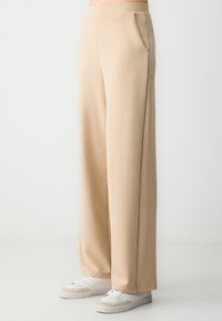 Beige wijde broek gemaakt van zachte stof, met zijzakken en contrasterende stiksels langs de naden. Gepaard met witte sneakers.