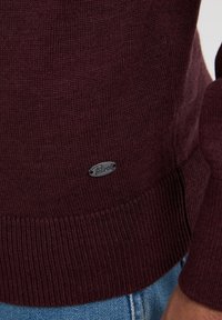Burgunderfarbener Strickpullover mit gerippten Bündchen und Saum, versehen mit einem silbernen Logotag, der auf der unteren linken Seite angenäht ist. Glatte Textur.