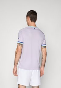 Homme avec les cheveux courts et foncés vue de dos, portant un t-shirt de sport léger violet avec des bordures bleues et un short blanc sur fond uni.