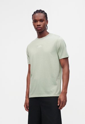 Jovem com cabelo trançado a usar uma t-shirt Oakley verde clara e calções pretos, em pé contra um fundo branco liso.