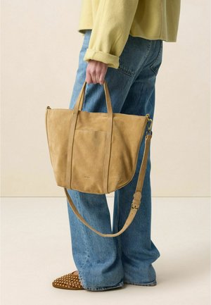 Personne portant un jean bleu et un pull jaune tenant un grand sac fourre-tout en daim beige avec une bandoulière, sur un fond uni.