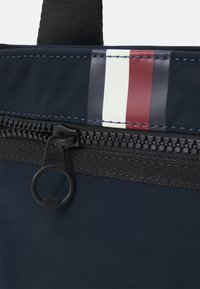 Borsa in nylon blu navy con zip nera, caratterizzata da un accento tessile a strisce in rosso, bianco e blu scuro. Manico superiore attaccato.