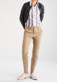 Beige housut, joissa on suloinen pinta, yhdistettynä raidalliseen napitettavaan paitaan ja mustaan bomber-takkiin. Valkoiset lenkkarit täydentävät ilmeen.