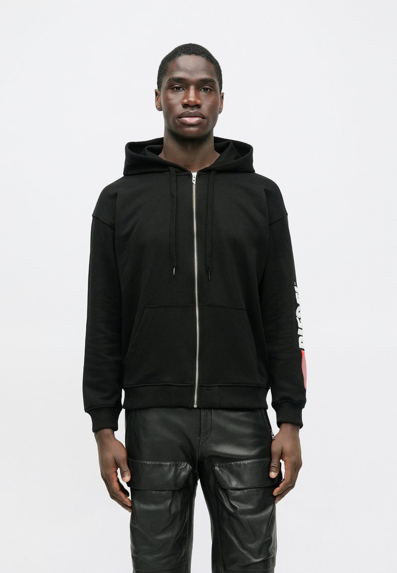 Muškarac u crnom zip-up dukserici s crvenim i bijelim tekstom na rukavu i crnim kožnim cargo hlačama, stoji nasuprot jednostavne bijele pozadine.