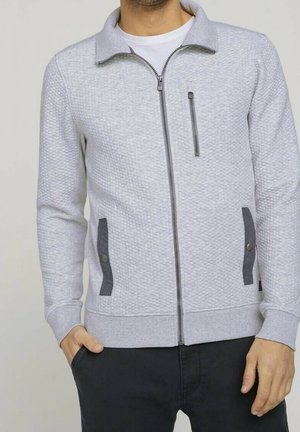 Homme portant une veste zippée texturée gris clair avec des accents gris foncé aux poches et un pantalon noir, debout avec une main dans la poche.