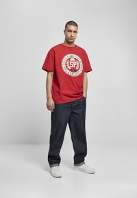 Tricou roșu din bumbac cu un imprimeu grafic alb în centrul său. O pereche de blugi largi din denim închis și adidași albi completează ținuta.