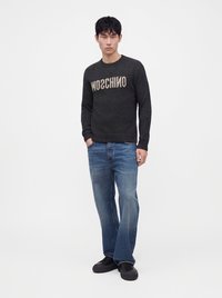 Jeune homme debout les mains dans les poches portant un pull noir avec un texte inversé, un jean large bleu et des chaussures noires.