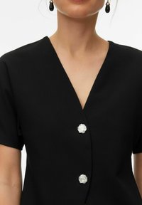 Mustast lühikeste varrukatega top sügava V-kaelusega ja hõbedaste kestakujuliste nuppudega. Ühtlane kangas minimalistliku disaini ja peene elegantsiga.