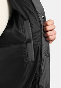 Hand die een open zwarte jas vasthoudt en de binnenzak met knoop toont, evenals de zichtbare ritssluiting en drukknopen bij de opening van de jas.