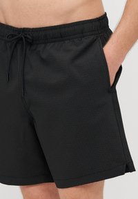 Sorte svømmebukser med teksturert overflate, elastisk midjebånd og snor. Lommer på siden med en liten splitt i kanten for fleksibilitet.