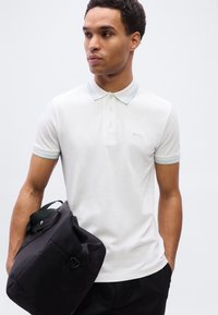 Λευκό πόλο με ανοιχτό μπλε γιακά, κοντά μανίκια και διακριτικό λογότυπο «BOSS». Το μοντέλο κρατάει μια μαύρη τσάντα, στέκεται μπροστά σε σκηνικό με απλό φόντο.