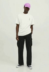 Homem com um boné lilás claro, t-shirt branca com um pequeno logótipo, calças cargo pretas e ténis pretos com solas brancas, posicionado contra um fundo simples.
