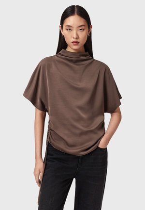 Kvinde med langt mørkt hår iført en løs brun satintop med korte ærmer og sorte bukser, der står mod en ensfarvet lys baggrund.