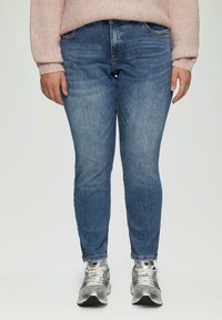 Vaqueros ajustados de mezclilla azul con una cintura de altura media, patrones de desgaste ligeros y cremalleras laterales en el dobladillo, combinados con zapatillas deportivas plateadas.