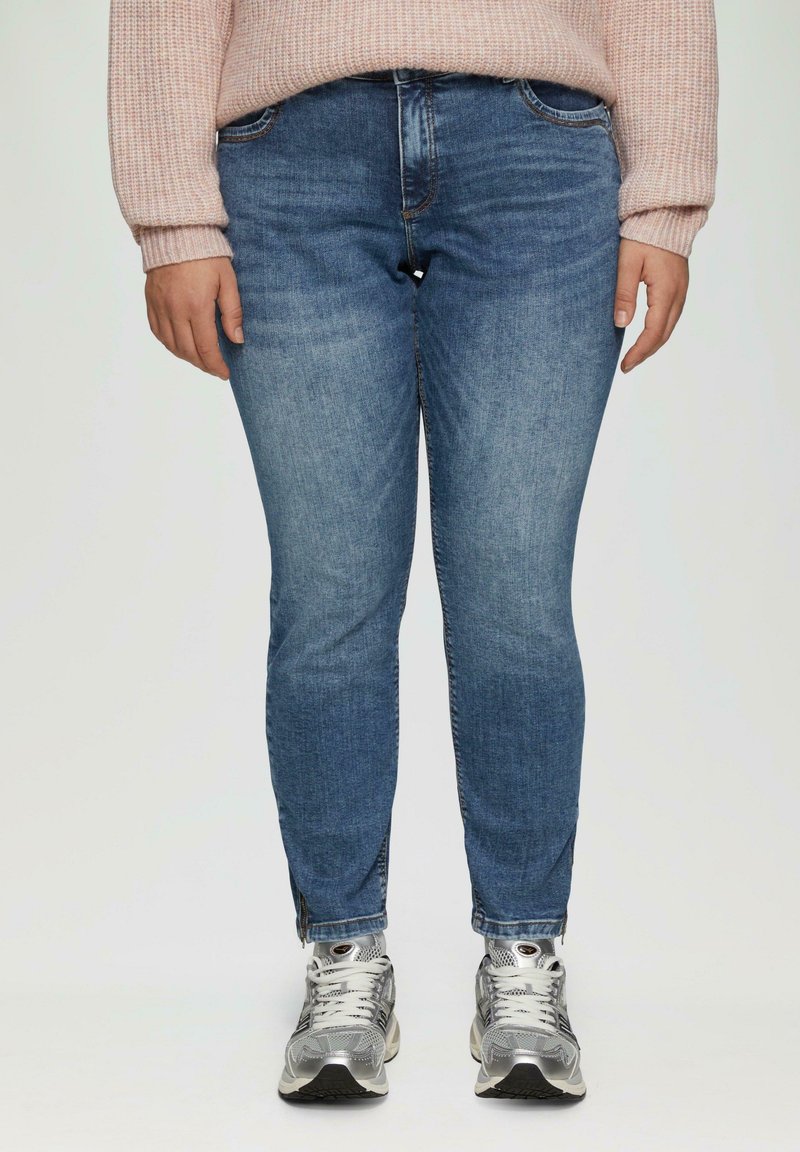 Vaqueros ajustados de mezclilla azul con una cintura de altura media, patrones de desgaste ligeros y cremalleras laterales en el dobladillo, combinados con zapatillas deportivas plateadas.