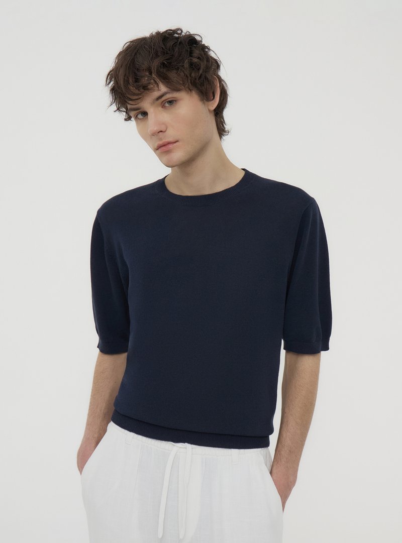 Maglione blu navy a maniche corte in un morbido materiale a maglia, con scollatura a girocollo e orlo aderente. Il modello lo indossa con pantaloni di colore chiaro.