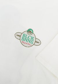 Witte katoen t-shirt met een ronde groen en beige logo met de tekst "BLCK BNNS" en de zin "Through the Desert" daaronder.