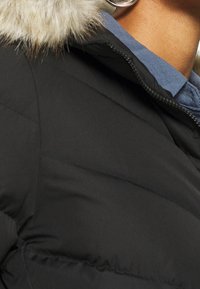 Zwarte pufferjack met een met bont gevoerde capuchon. Heeft een geblokt patroon en een ritsfront. Gelaagd met een blauwe stof eronder.