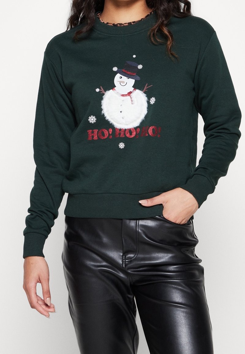 Sweatshirt vert foncé avec un graphisme de bonhomme de neige portant un chapeau noir, une écharpe rouge, et le texte « HO ! HO ! HO ! » en paillettes rouges ; poignets et ourlet côtelés.