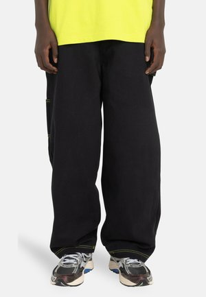 Loose black cotton pants with side pockets, contrasting yellow stitching, paired with multi-colored athletic shoes featuring mesh details.