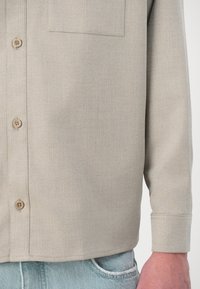 Camicia beige con bottoni, motivo a spina di pesce, taschino sul petto e maniche con polsini. Il materiale sembra liscio con una vestibilità strutturata.