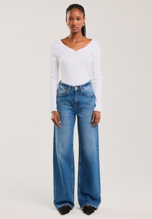 Jauna sieviete ar bizēm, ģērbusies baltā garroku V veida kakla topā un augsta vidukļa platām zilām džinsām, stāv pret vienkāršu fonu.