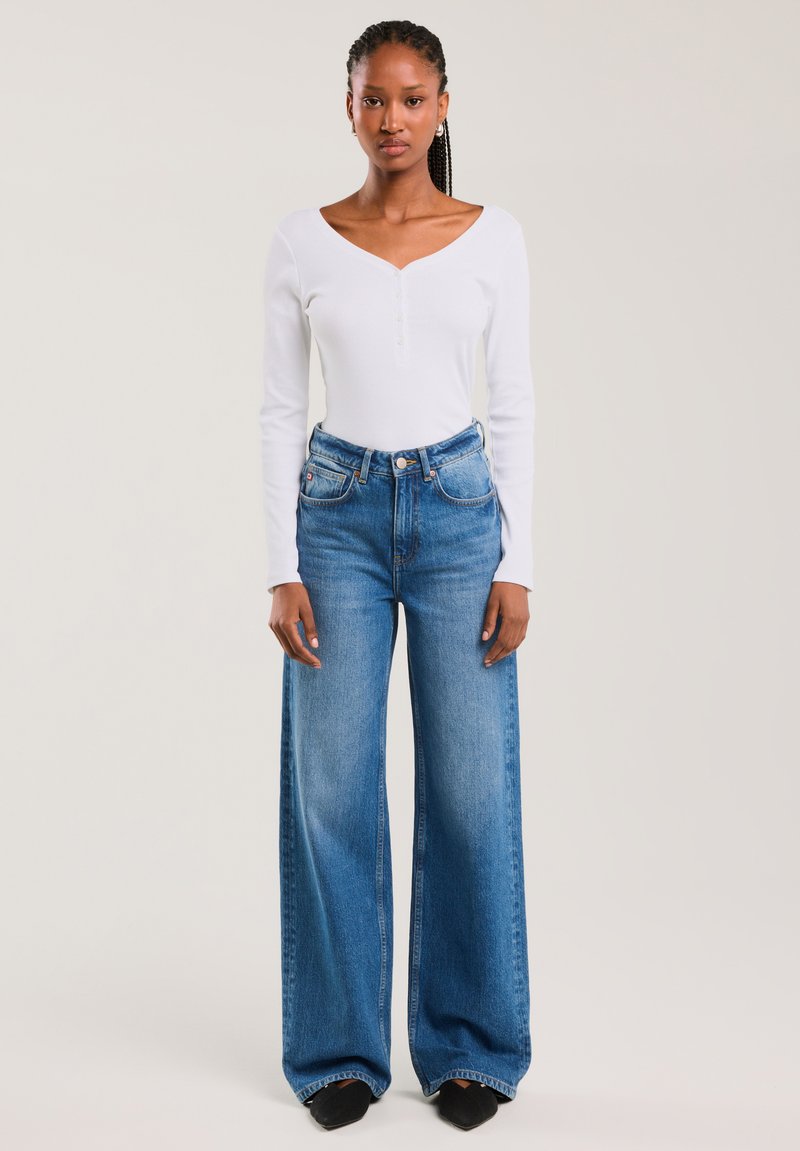 Jeune femme avec des tresses portant un haut blanc à manches longues et col en V, ainsi qu'un jean bleu taille haute à jambes larges, debout contre un fond uni.