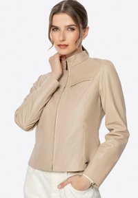 Veste en cuir synthétique beige avec un col haut, fermeture éclair à l'avant et deux poches latérales. Présente un design élégant et ajusté avec un léger éclat.