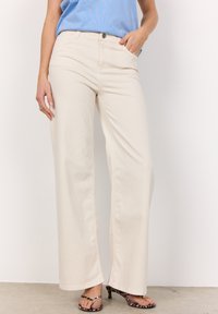 Beige vida jeans i bomull med framfickor, knappstängning och synlig sömdetalj. Bärs med en blå topp och mönstrade klackar.