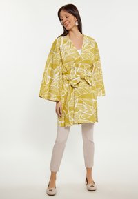 Keltainen kimono-tyylinen takki, jossa on valkoisia lehtikuvioita, vyötetty vyötäröltä, yhdistettynä vaaleanbeigeihin housuihin ja vaaleisiin liukuhihnakenkiin.