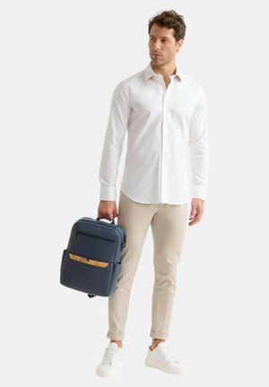 Zaino blu navy dal design elegante, con una tasca frontale con un accento in legno, indossato da una persona con una camicia bianca e pantaloni beige.