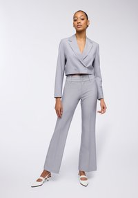 Blazer grigio a vita corta con doppi bottoni, abbinato a pantaloni a gamba larga coordinati. La modella indossa tacchi a punta bianchi. Tessuto liscio e sartoriale.