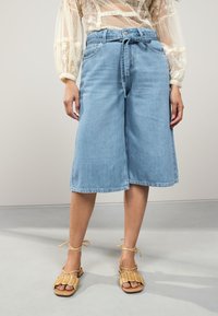 Világoskék farmer culottes övvel a derékrészen, széles szárakkal és nyers szegéllyel, bézs színű, fonott szandállal párosítva.