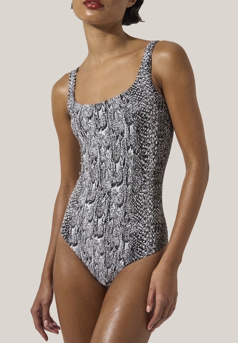 Kvinna i en svart och vit ormtryckt ärmlös enstycks baddräkt, med armarna avslappnat längs sidorna, mot en enkel ljus bakgrund.