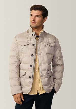 Beige pufferjack met een kraag, vier voorzakken en een knoopsluiting. Vriendelijke textuur en subtiele glans, gedragen over een gele shirt.