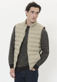 Beige vadderad väst med hög krage, dragkedja och sidofickor, lagd över en strukturerad mörk tröja med blå och gyllene ränder.