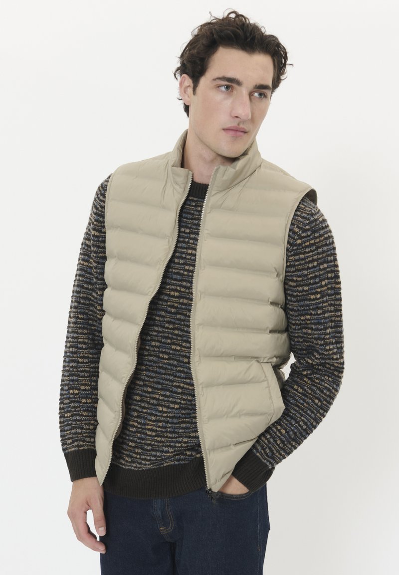 Beige vadderad väst med hög krage, dragkedja och sidofickor, lagd över en strukturerad mörk tröja med blå och gyllene ränder.