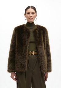 Brown faux fur jacket with a cropped silhouette, rounded neckline, and soft texture; paired with a green high-neck top and brown belt.