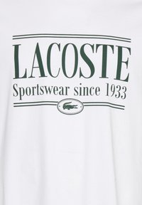 Bílý bavlněný tričko se zeleným textem: "LACOSTE Sportswear od roku 1933," s malým krokodýlím logem v oválu pod textem.