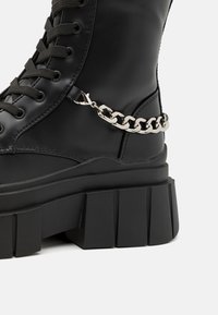 Μαύρη δερμάτινη μποτάκι αστραγάλου με χοντρή σόλα, που διαθέτει κορδόνια στο μπροστινό μέρος και κομψή ασημένια αλυσίδα στη πλευρά.