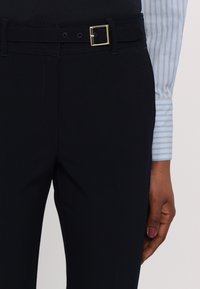 Gros plan d'une personne portant un pantalon noir sur mesure avec une ceinture assortie et une manche de chemise rayée bleu clair visible.