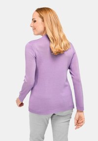 Frau mit schulterlangen blonden Haaren, die einen langärmeligen lila Pullover und hellgraue Hose trägt, von hinten gezeigt.