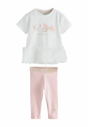 Ung pige med kort sort hår iført en hvid kjole med blomsterdetaljer, pink leggings og hvide sneakers, stående på en grå gulv.