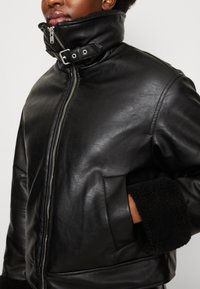 Giacca in pelle nera con colletto alto, zip argentata e polsini foderati in pelliccia. Presenta tasche laterali e una texture liscia e lucida.